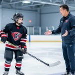 Youth hockey player in full gear standing on an indoor ice rink with a coach offering encouragement, with a blurred team setting in the background.