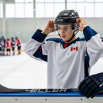 Young Canadian junior hockey player adjusting his helmet at an indoor rink, with hockey gear and blurred teammates in the background.