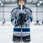 Youth hockey player holding a properly sized hockey stick on an indoor ice rink, with sharp focus on the stick grip and blade.
