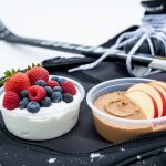 Close-up of Greek yogurt with berries and nut butter with sliced apples packed beside a hockey gear bag with skate and stick equipment in the background.